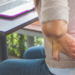 woman sitting at desk working on a computer with hands pressed on her lower back indicating back pain