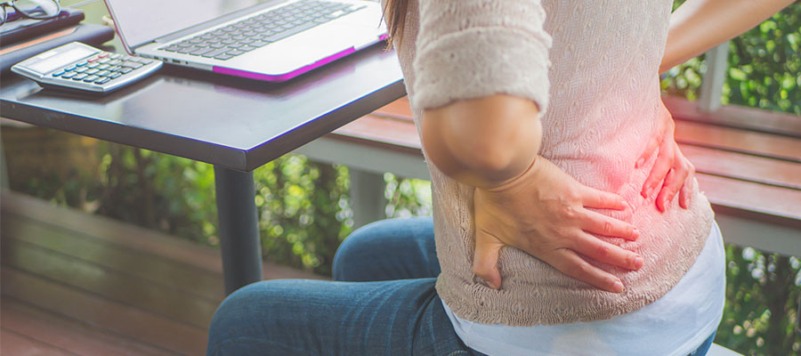 woman sitting at desk working on a computer with hands pressed on her lower back indicating back pain